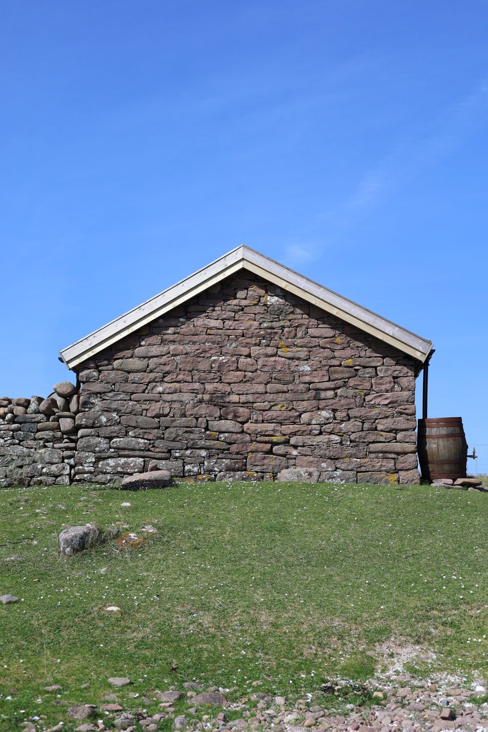 Rustic stone building with pitched roof sits on grassy hill against bright blue sky, with wooden barrel beside it.