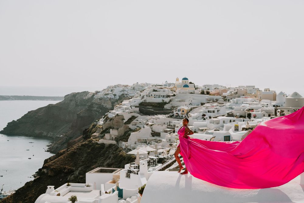 A bright pink flowing dress creates dramatic movement against the coastal architecture of Santorini.
