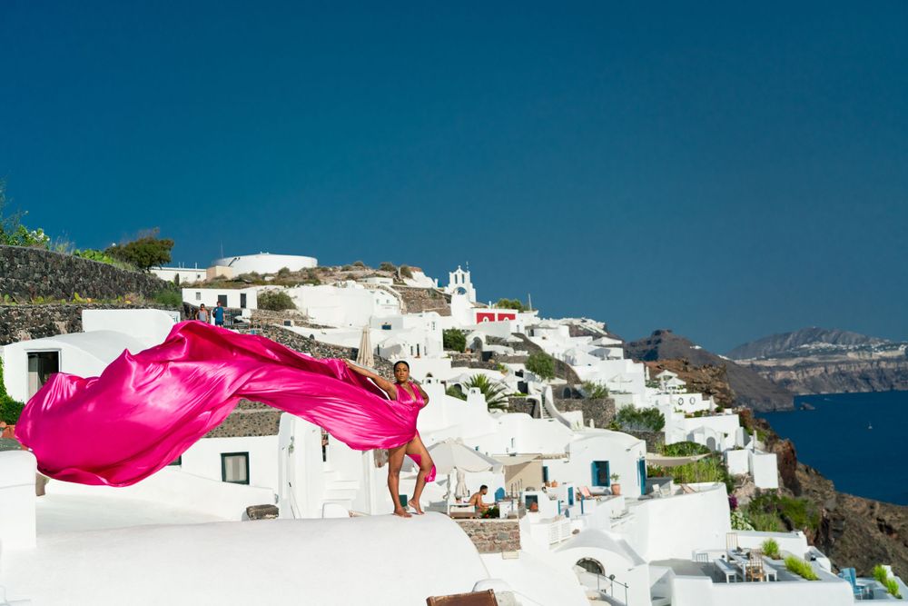 A bright pink dress billows in the wind against white buildings and blue seas of Santorini.