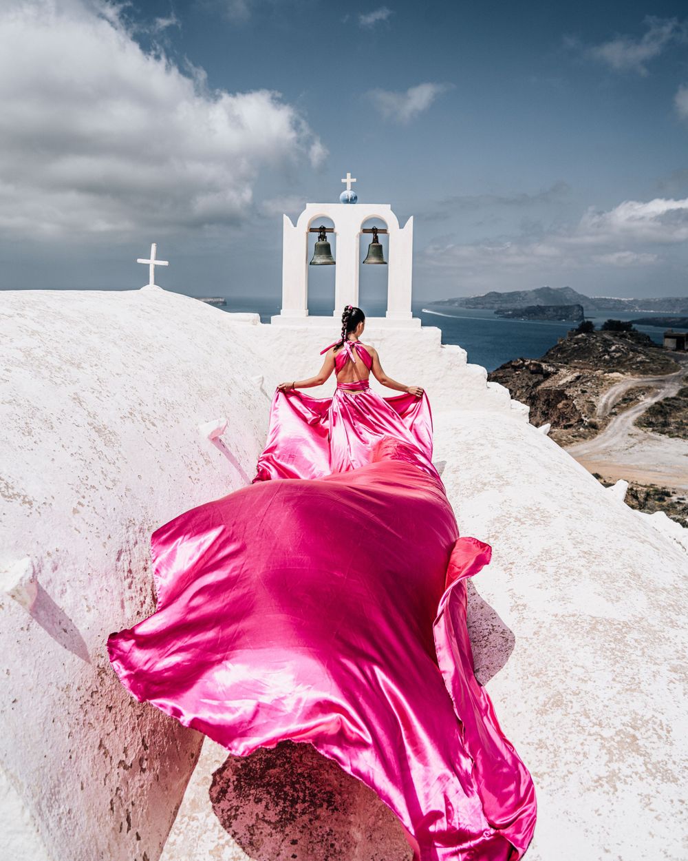 A flowing pink dress frames a white church bell tower overlooking the Aegean Sea in Santorini.