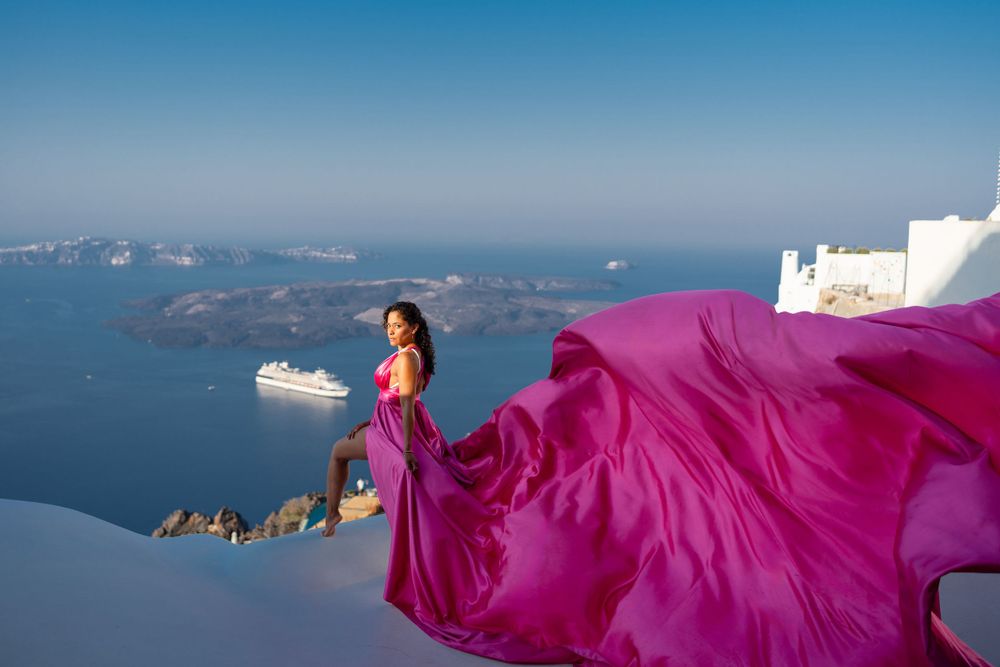 A flowing magenta dress catches the wind on a Santorini rooftop with cruise ship in background.