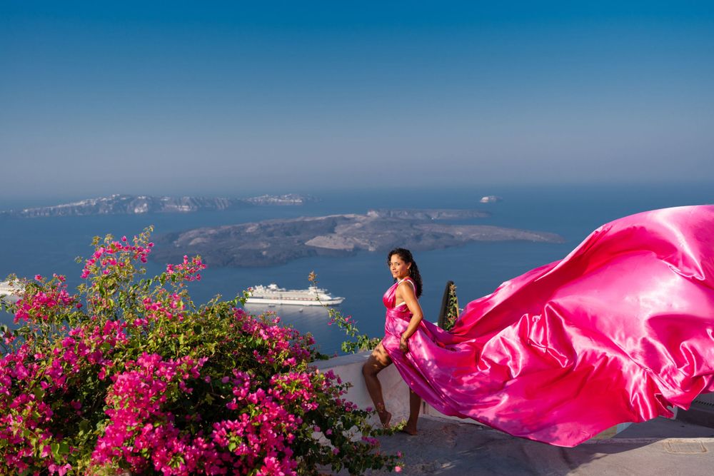 A billowing pink dress frames Santorini's iconic landscape with bougainvillea and sea views.