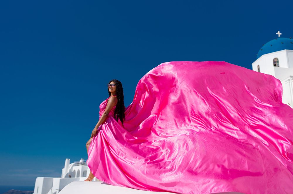 Flowing hot pink dress billows dramatically against white buildings and blue sky in Santorini, Greece.