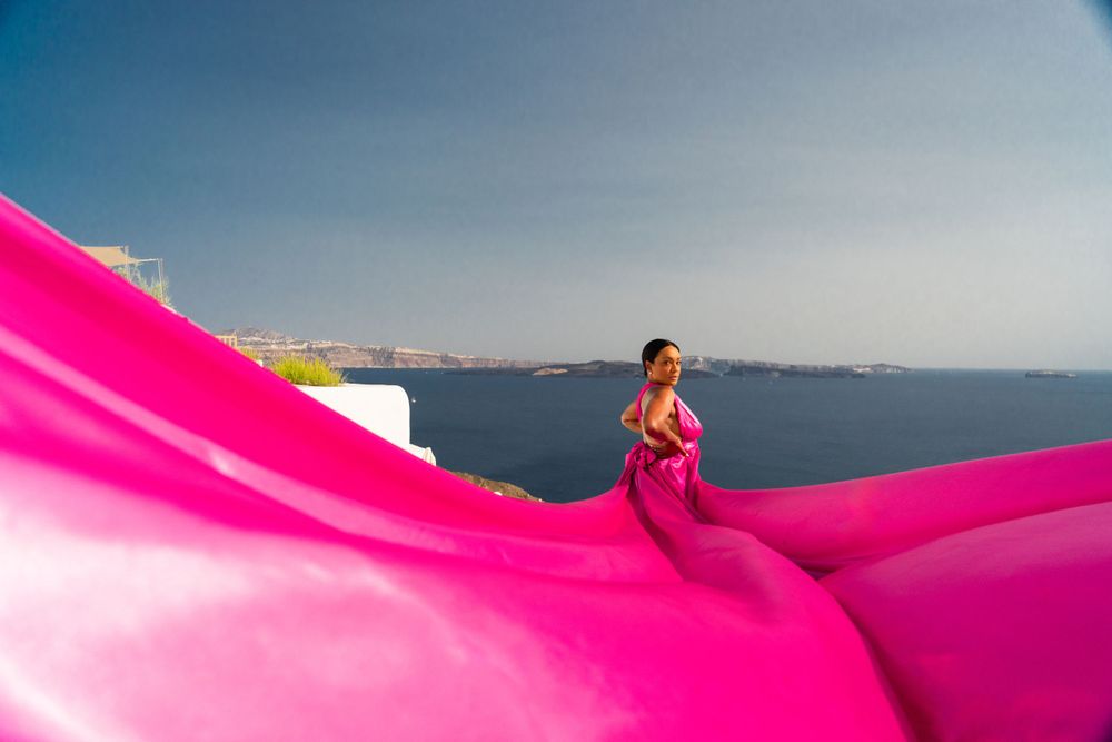 Bright pink flowing dress creates dramatic silhouette against Santorini's coastal backdrop.