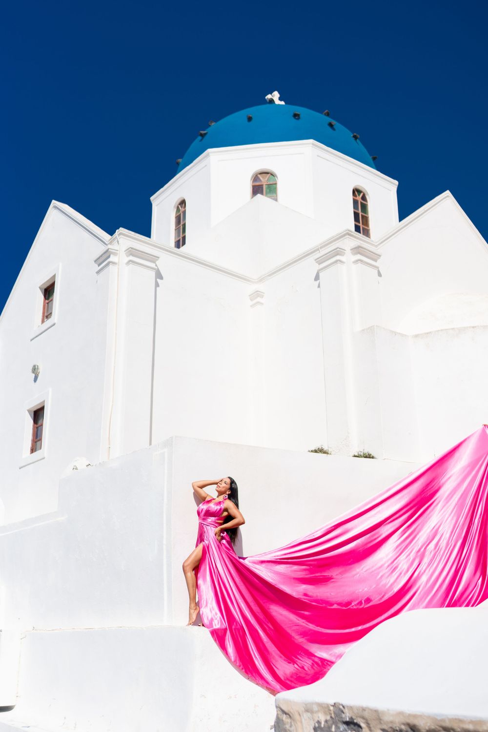 Flowing pink fabric contrasts against white church with blue dome in Santorini.