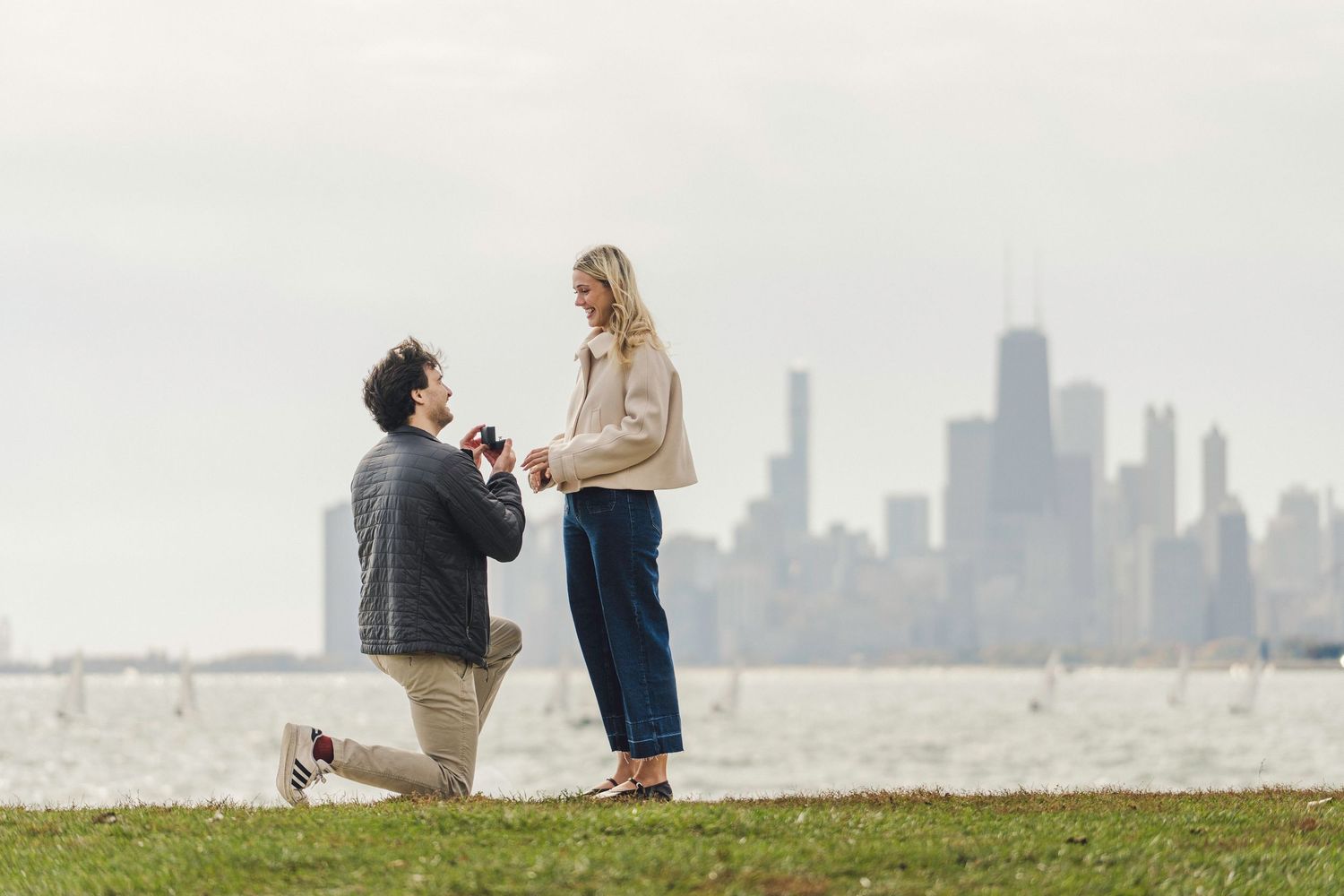 A man kneeling on one knee proposes to a smiling woman on a grassy lakefront with a hazy city skyline across the water in the background.