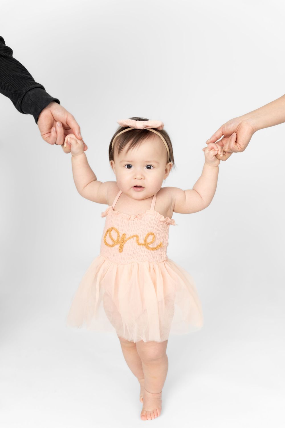 A baby wearing a peach-colored romper holds hands with adults while learning to walk in a bright studio setting.