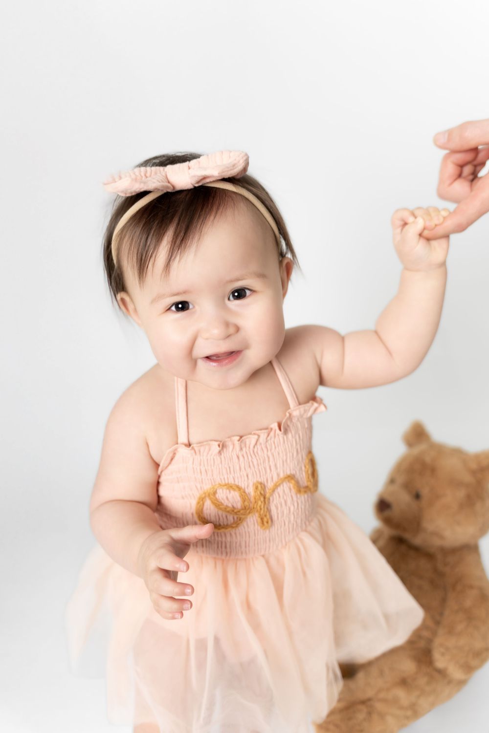 A baby in a peach-colored dress with a matching bow headband smiles and raises their arms while standing next to a teddy bear.