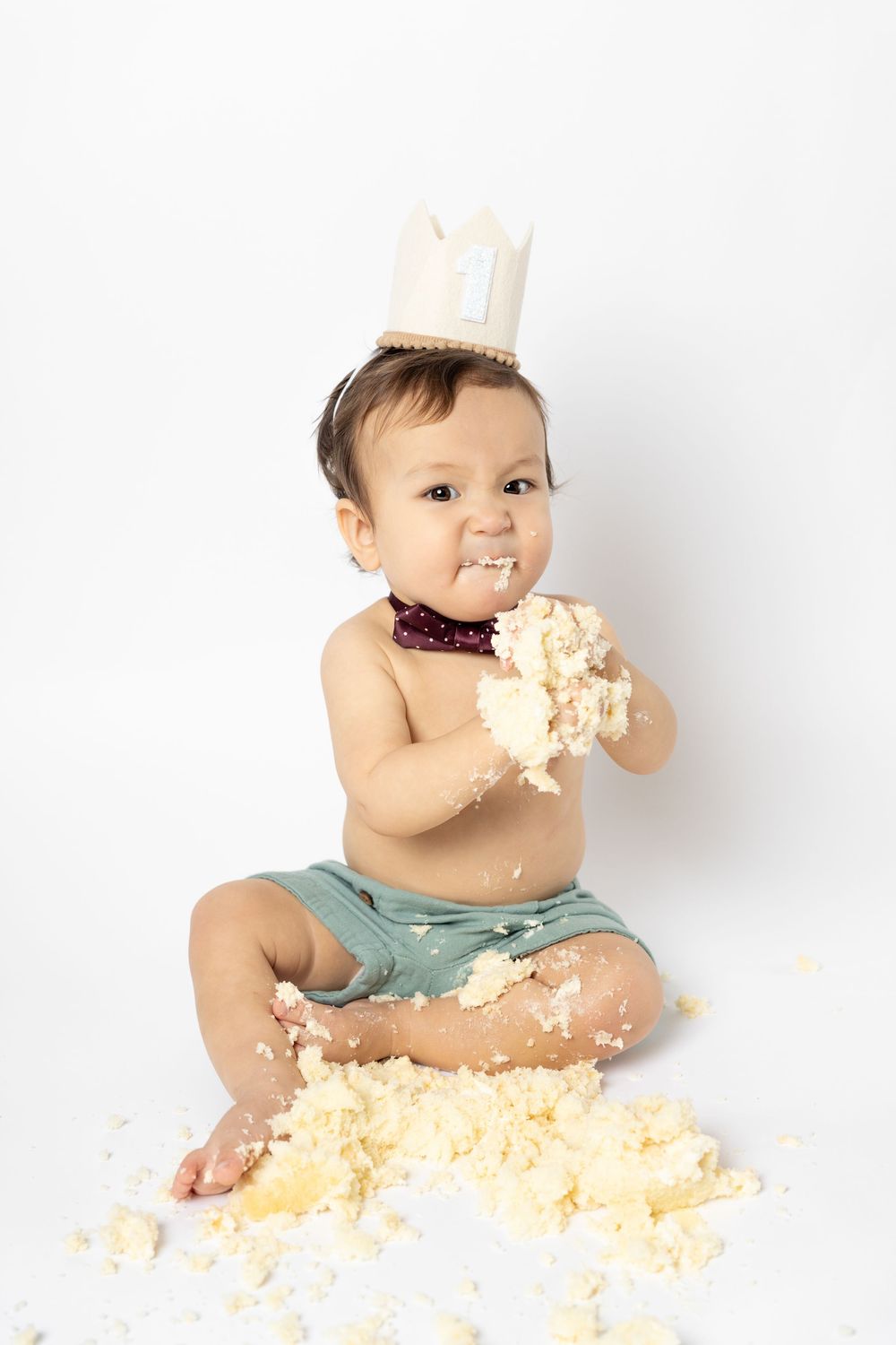 Baby in blue outfit plays with cake smash during first birthday photoshoot on white background.
