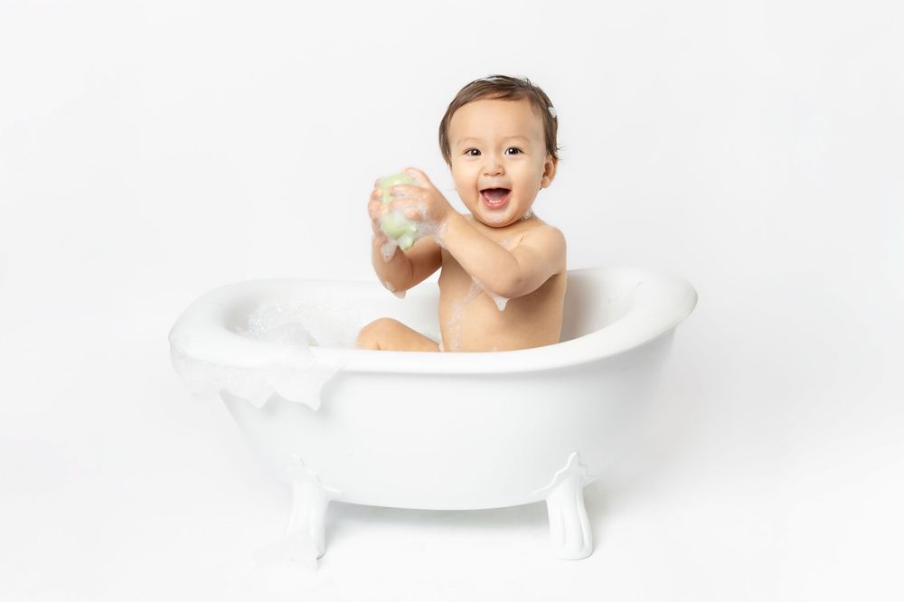Happy baby splashing and playing in a white bathtub against white background.