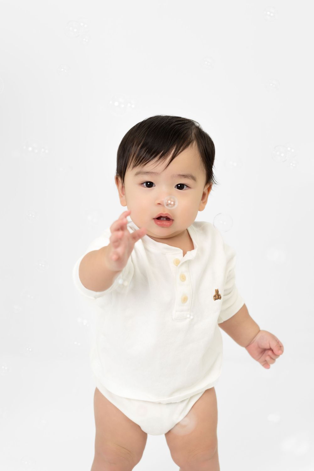 Baby wearing white polo onesie reaches out while standing against white background.