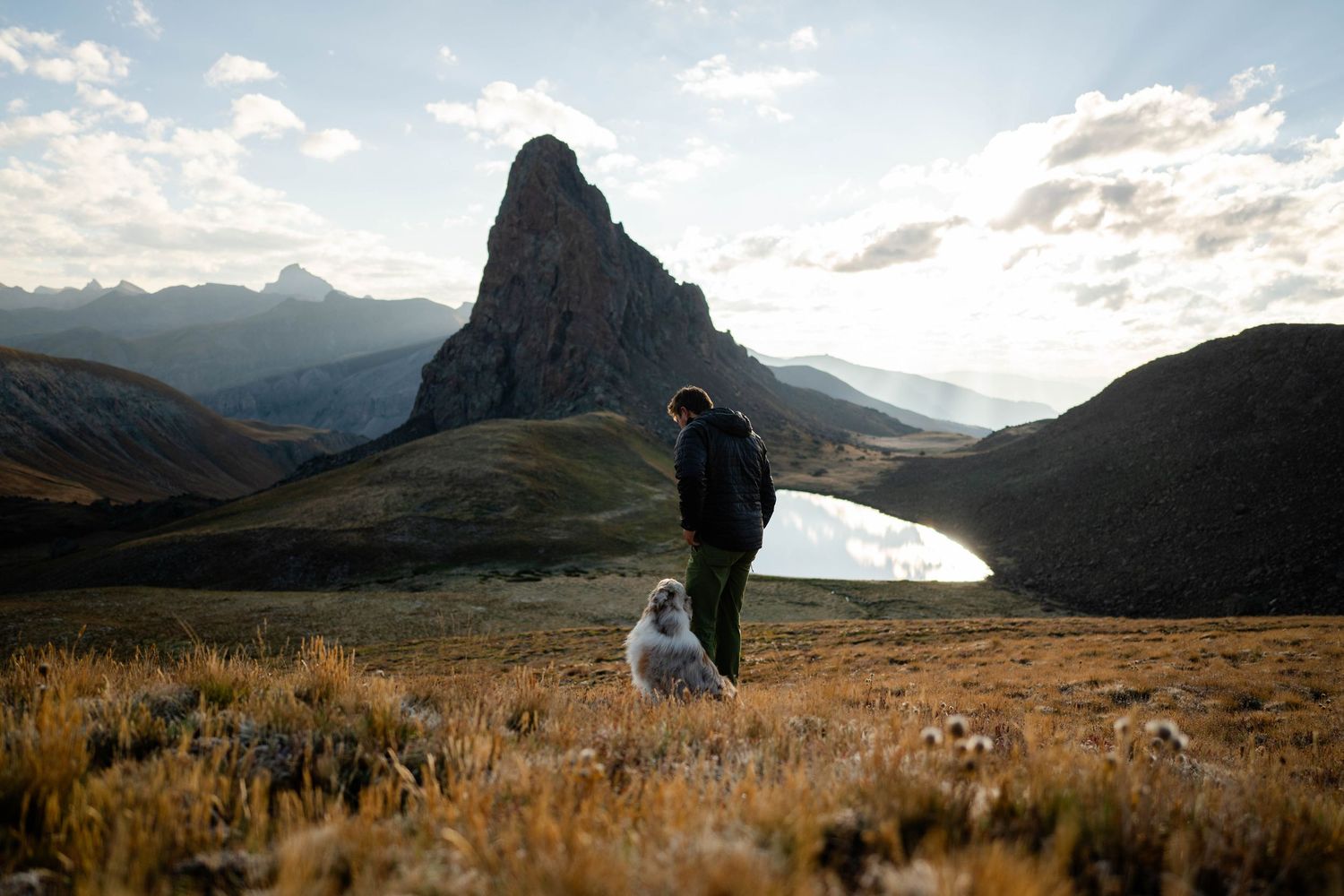 A panoramic mountain landscape at sunrise with man and his Australian Shepherd leaning against his leg in the San Juan Mountains of Colorado