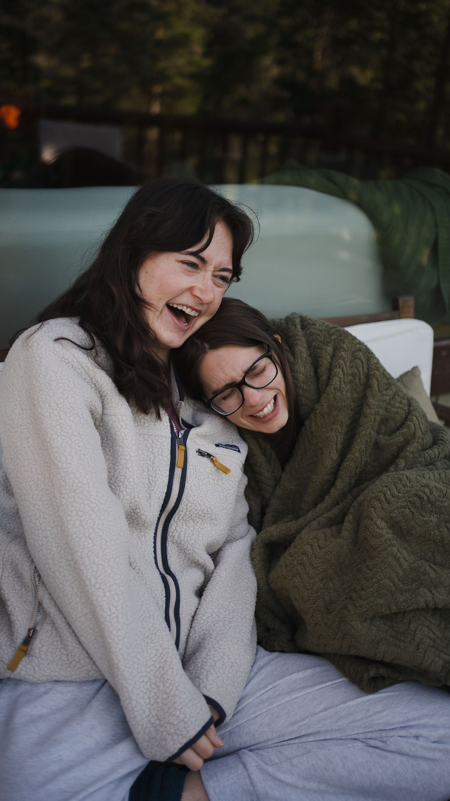 A portrait of two friends sharing a cozy laugh while wrapped in blankets outdoors.