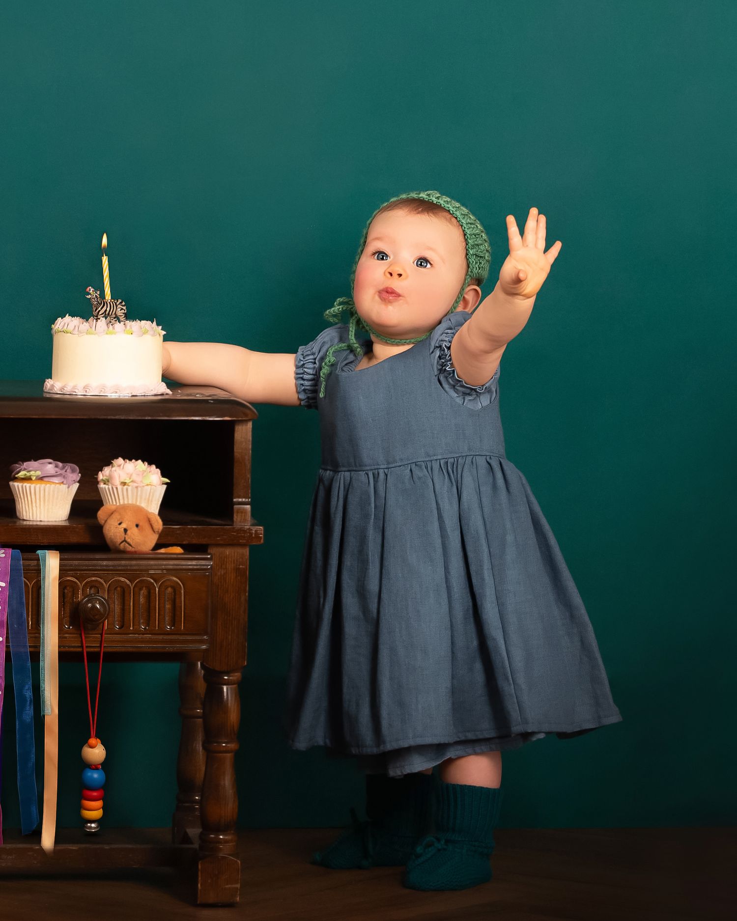 A young child wearing a blue dress holds up three fingers while standing beside a wooden shelf with a birthday cake. Birthday Heirloom