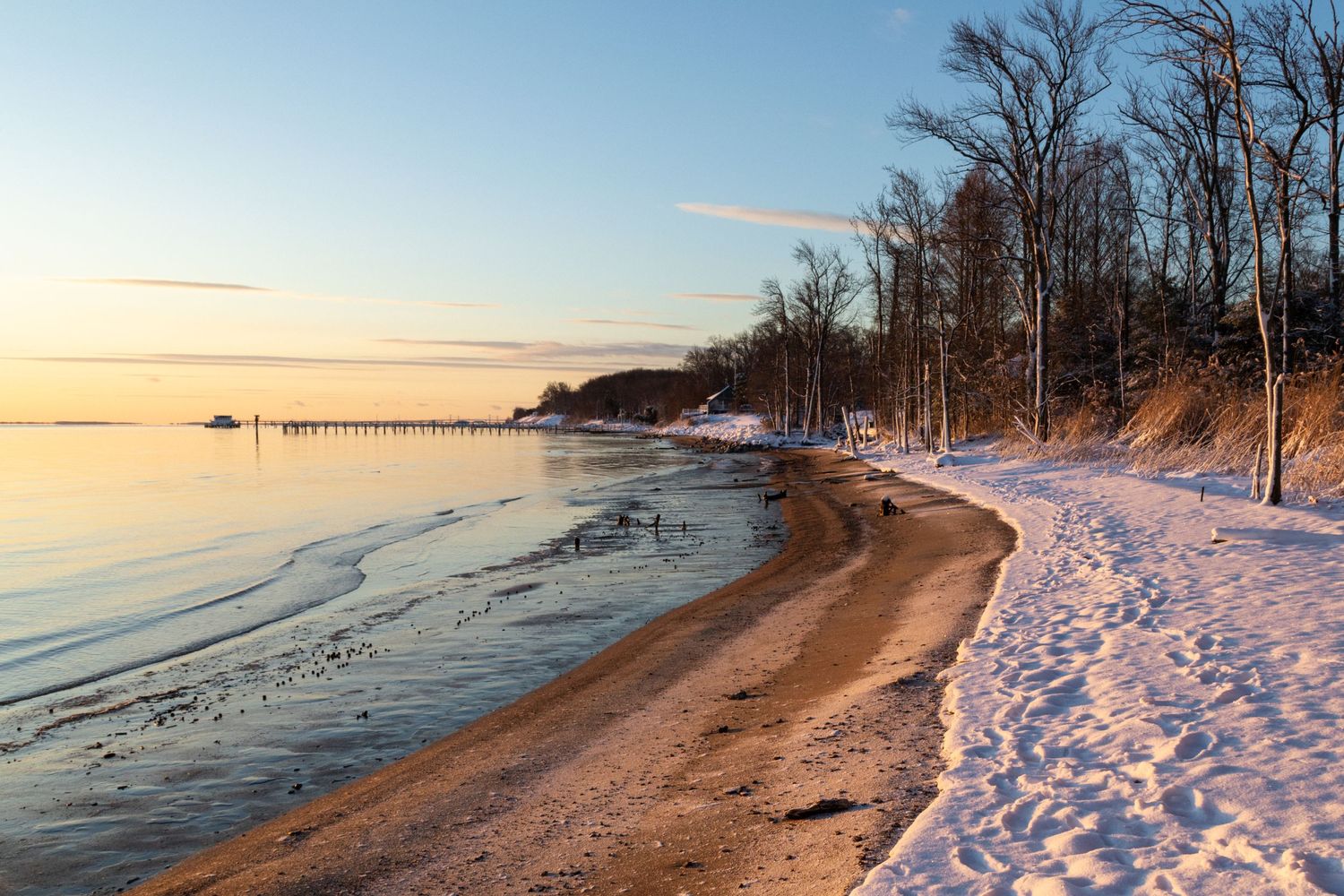 A serene sandy beach at a park is dusted with a layer of fresh snow, creating a striking contrast between the white snow and the golden sand