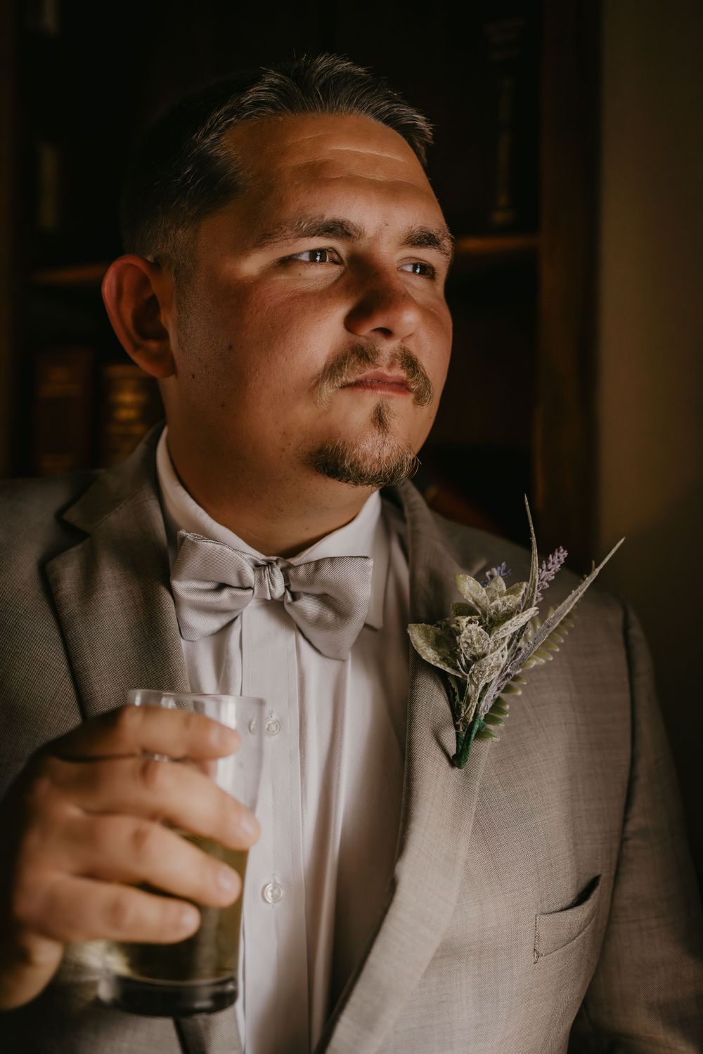 Groom in gray suit and bow tie holding a glass drink, wearing floral boutonniere in dim light.