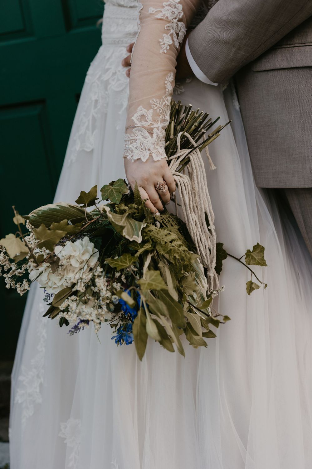 Bride in lace wedding gown holding rustic dried floral bouquet with greenery against teal door.