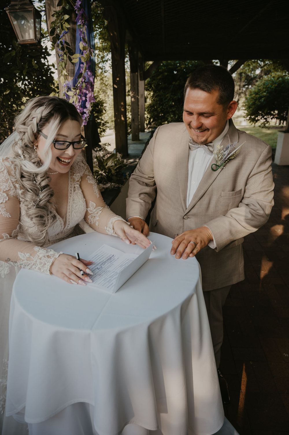 Smiling bride and groom signing wedding marriage certificate at outdoor ceremony.