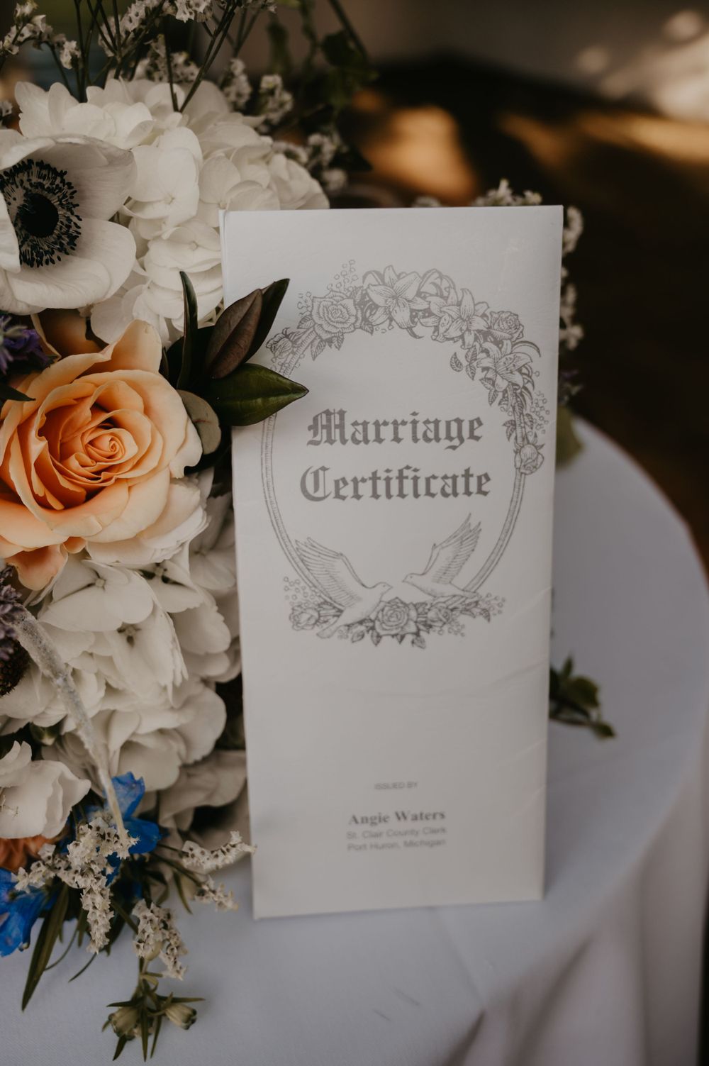 Elegant marriage certificate displayed beside peach roses and white anemone wedding flowers on white table.