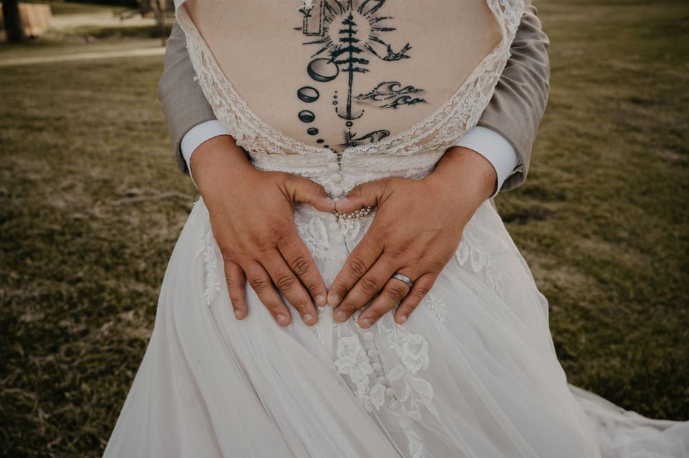 Couple forms heart shape with hands over brides lace wedding gown revealing intricate tattoo on torso.