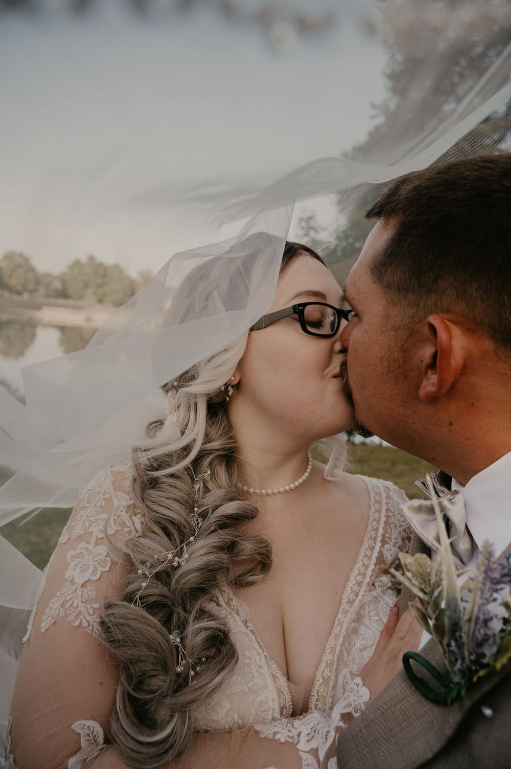 Bride wearing glasses and pearl necklace shares a kiss with groom under flowing white wedding veil.