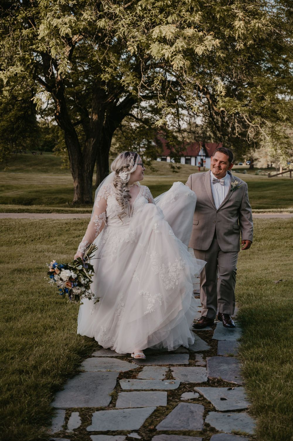 Bride in lace gown holding bouquet walks with groom in gray suit on stone path surrounded by lush greenery.