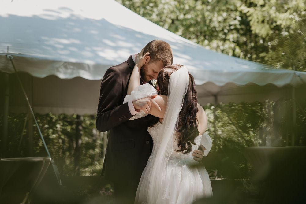 A bride and groom share a romantic kiss under a white tent canopy surrounded by lush green trees on their wedding day.