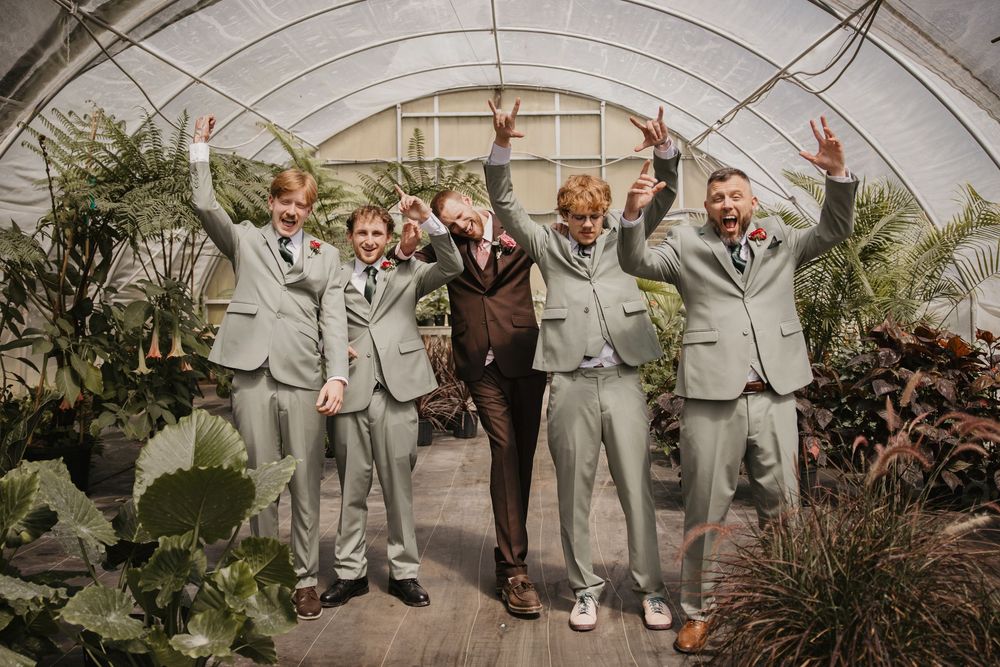 Groomsmen in sage green suits celebrate with groom in brown suit inside a lush greenhouse wedding venue.