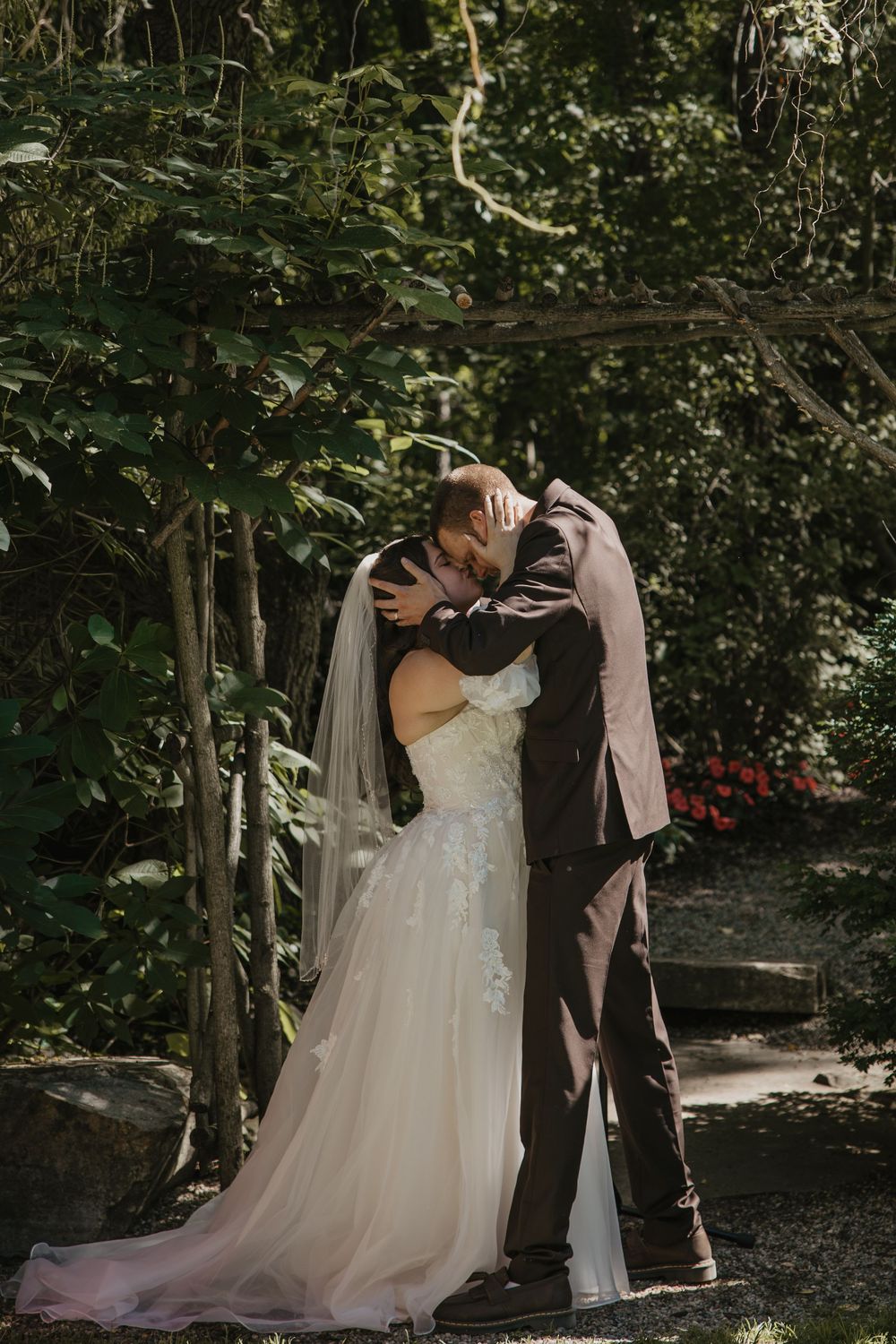 Bride and groom share a romantic first kiss under a rustic wooden arch surrounded by lush green garden foliage.