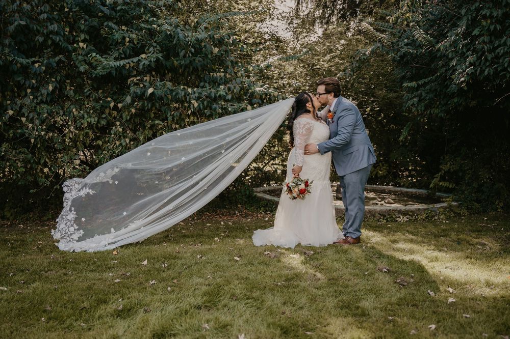 Bride and groom kissing outdoors as long cathedral veil dramatically blows in the wind behind them.