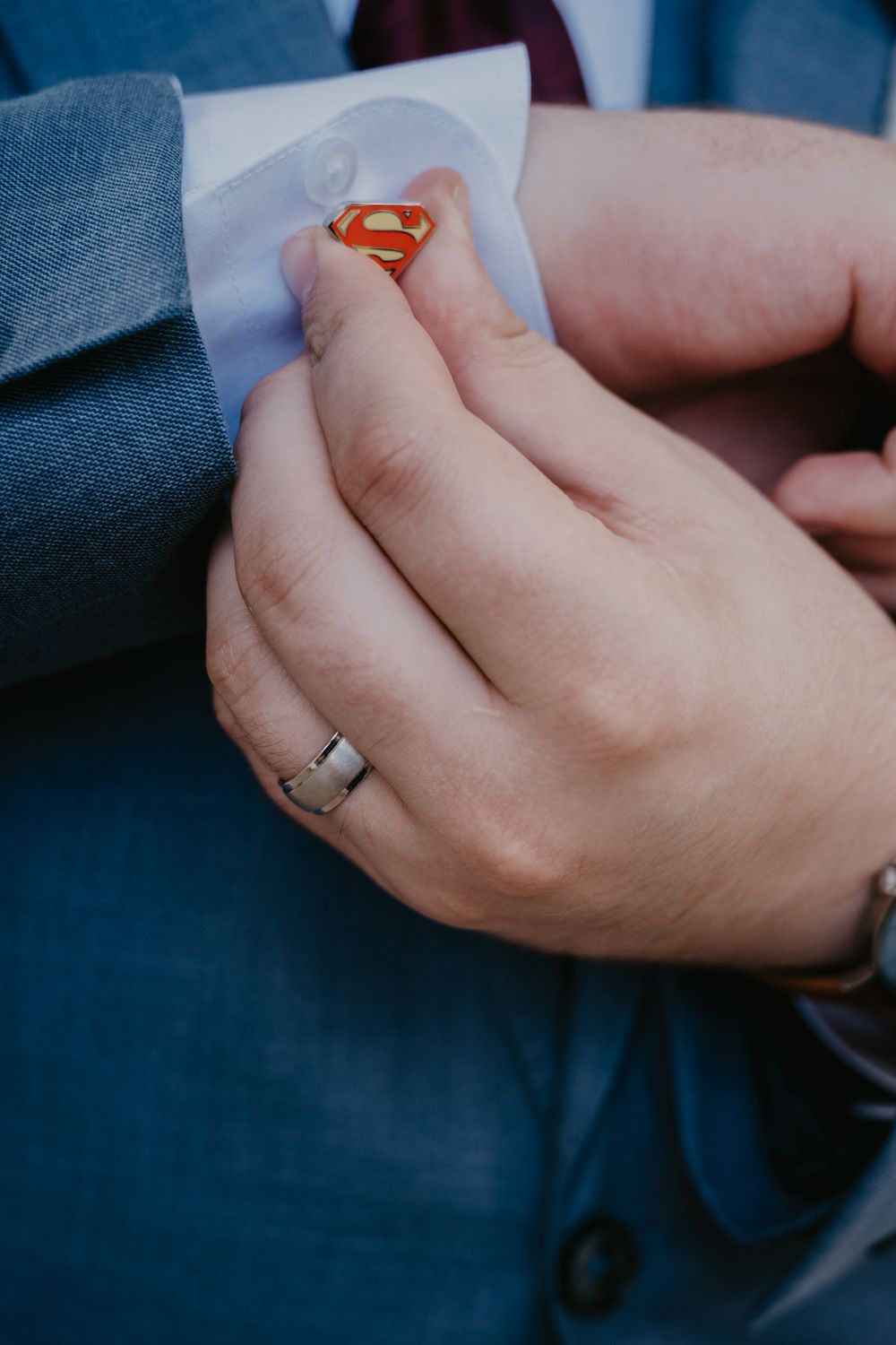 Close-up of groom's Superman cufflink on blue suit sleeve with wedding band on hand.