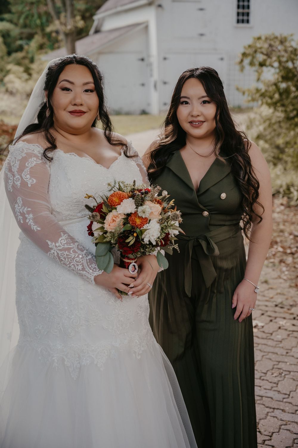 Bride in lace long sleeve gown holds autumn bouquet standing with bridesmaid in olive green jumpsuit.