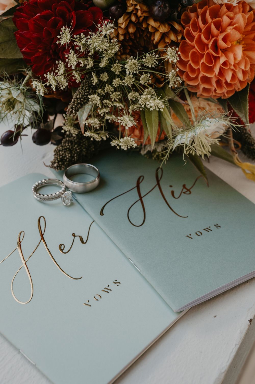 Her and His vows booklets with wedding rings and autumn floral bouquet featuring dahlias and berries.