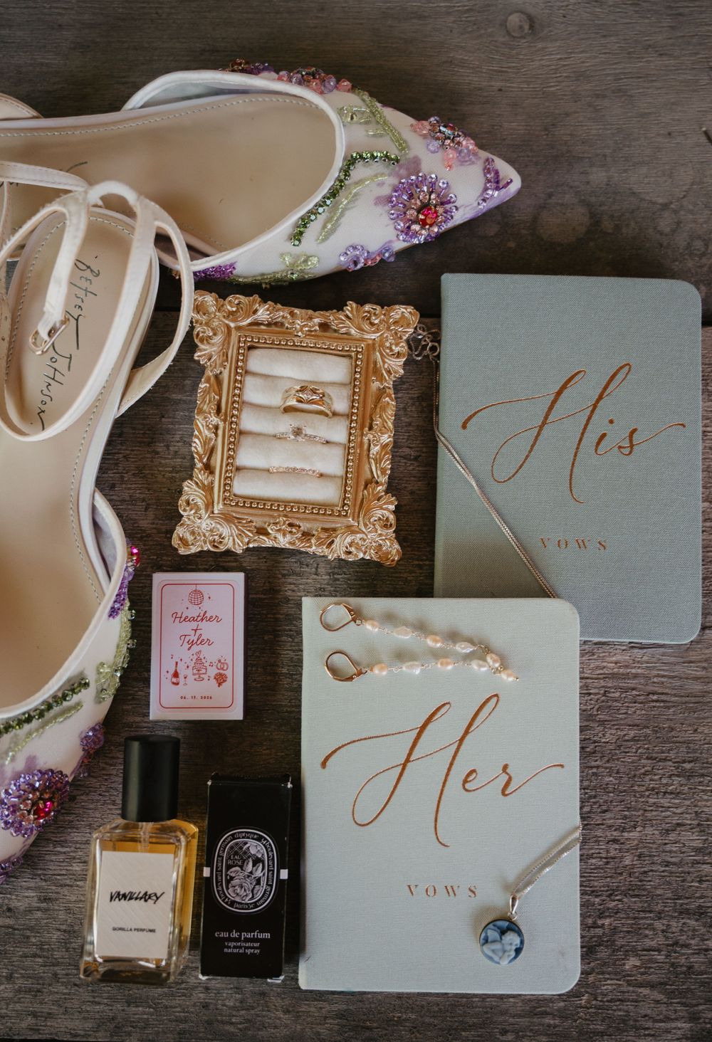 Elegant wedding flat lay featuring floral heels, gold ring frame, His and Her vow books, perfume bottles, and pearl jewelry on wood.