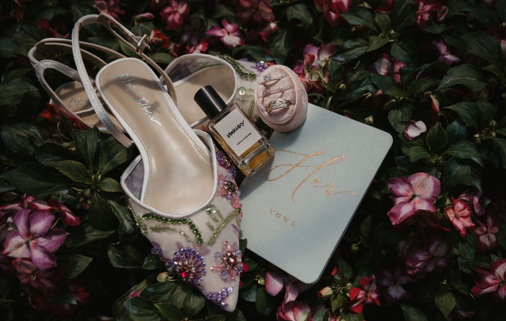 Bridal accessories flat lay with floral heels, perfume bottle, and blue card on pink flower bed.