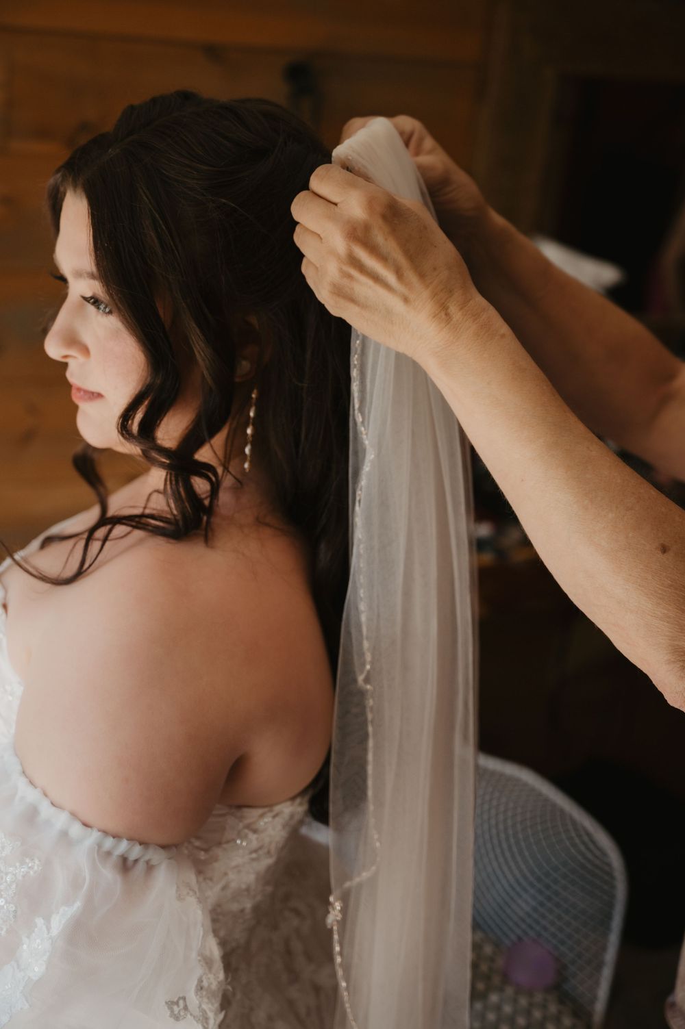 A bride in a white lace gown has her wedding veil pinned into her dark hair by a helper in a warm wooden room.