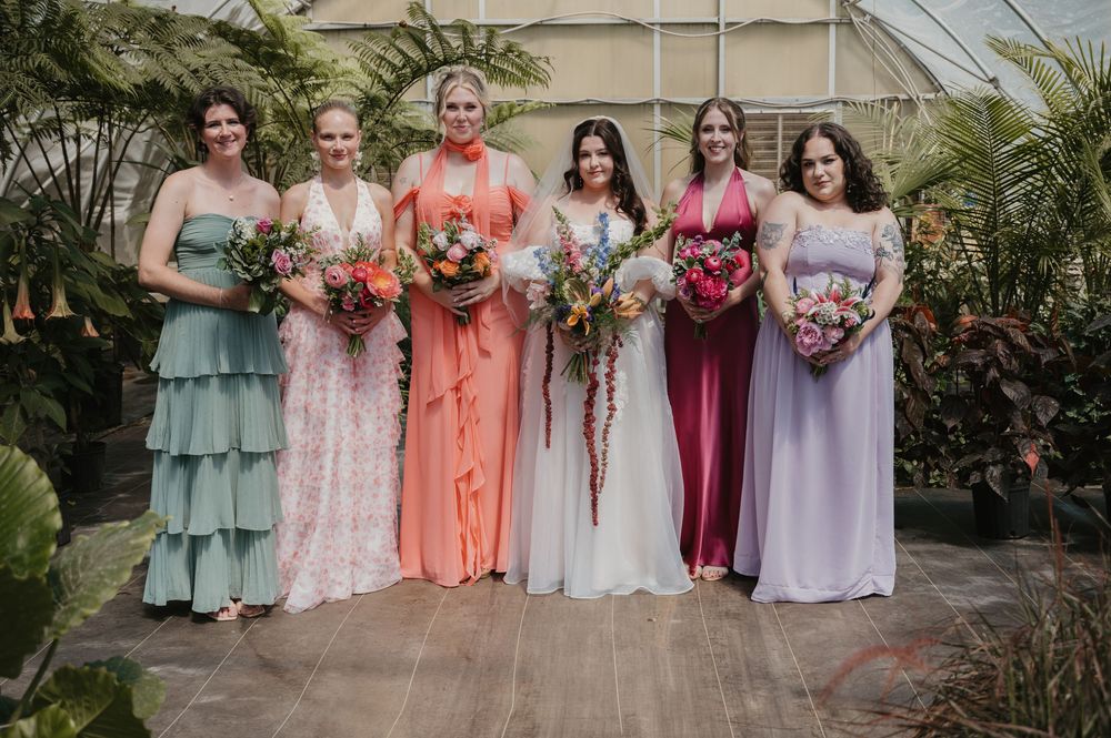 Six bridesmaids in colorful mismatched gowns holding floral bouquets pose together in a lush greenhouse wedding venue.