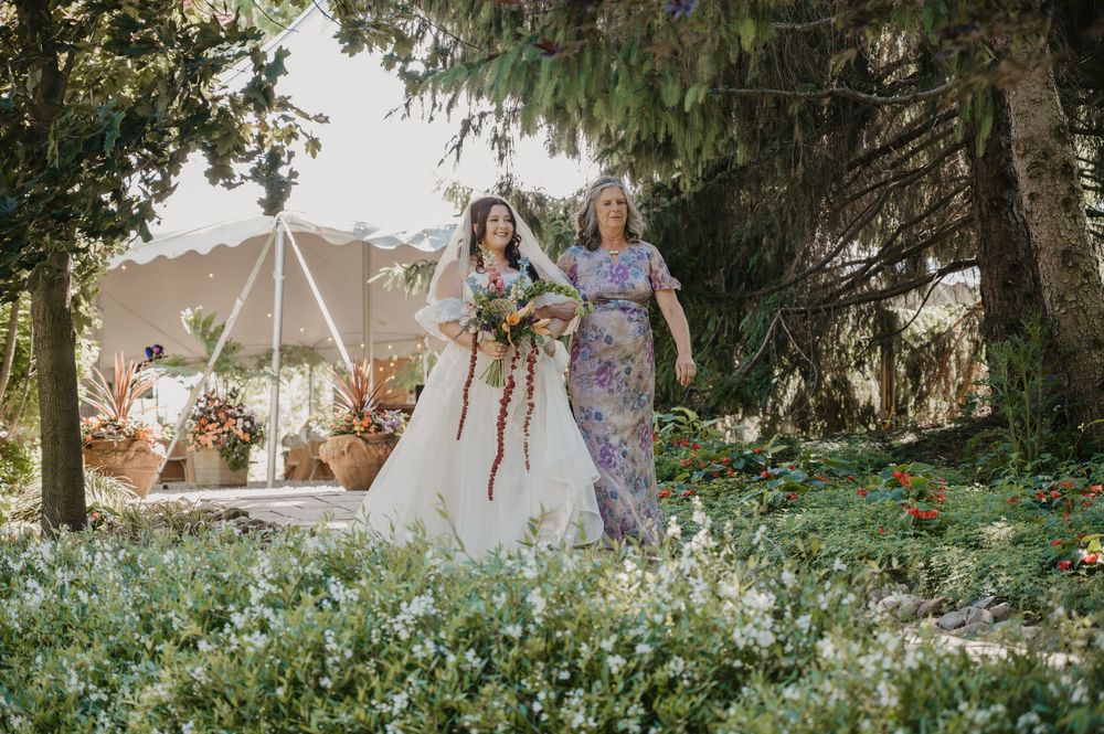 Bride in white gown walks with bridesmaid in floral dress through lush garden holding colorful bouquet.