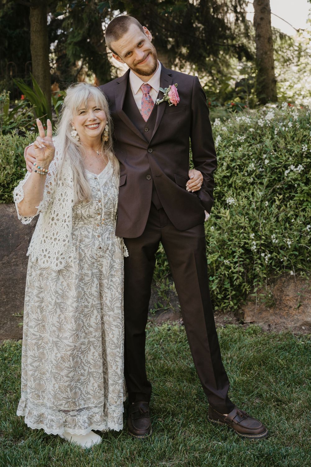 An older woman in a lace dress flashing a peace sign poses with a young groom in a dark suit at an outdoor wedding.