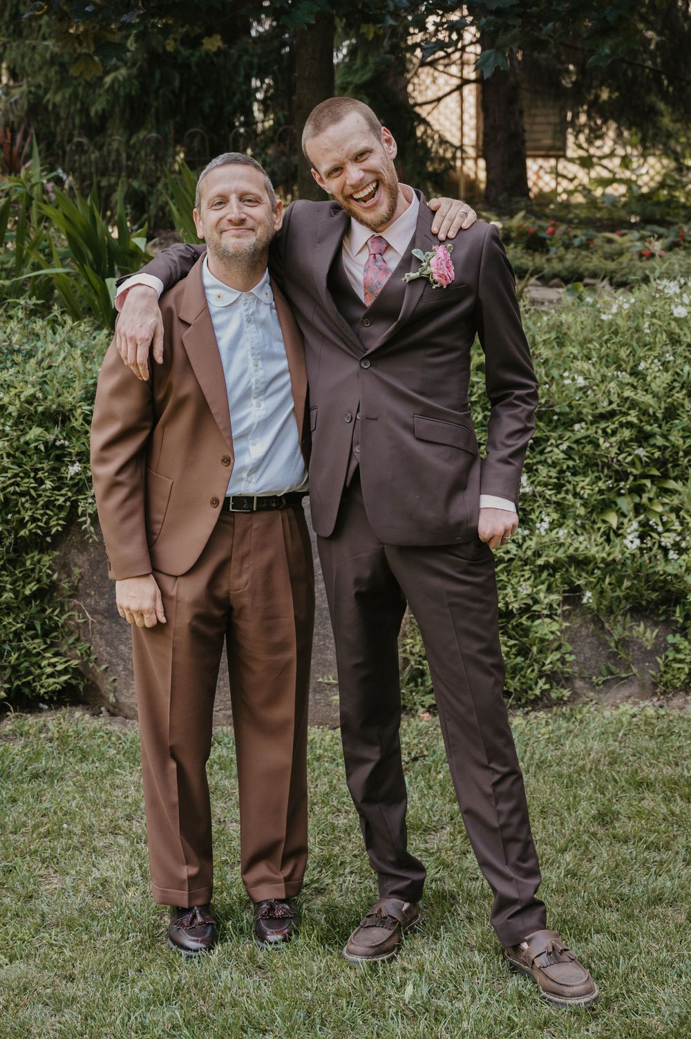 Two men in brown suits smile and pose together outdoors at a wedding, one wearing a pink boutonniere.