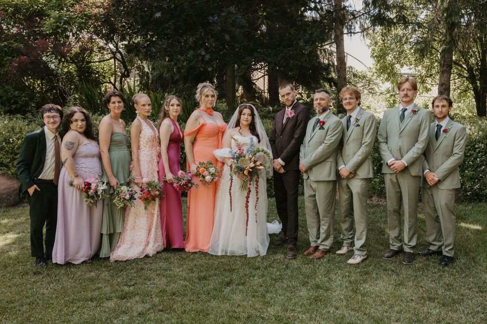 Wedding party posing outdoors on green lawn, bridesmaids in pink and sage gowns, groomsmen in tan suits flanking bride and groom.