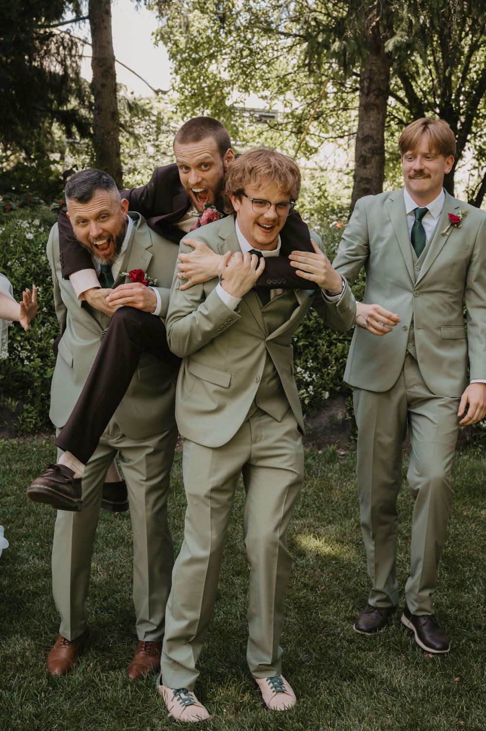 Groomsmen in sage green suits laughing and goofing around outdoors, one giving another a piggyback ride at a wedding.