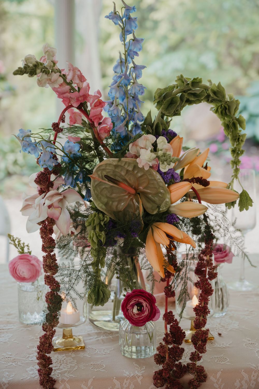 Colorful wedding floral centerpiece with lilies, delphiniums, anthurium, and ranunculus in glass vases on a lace tablecloth.