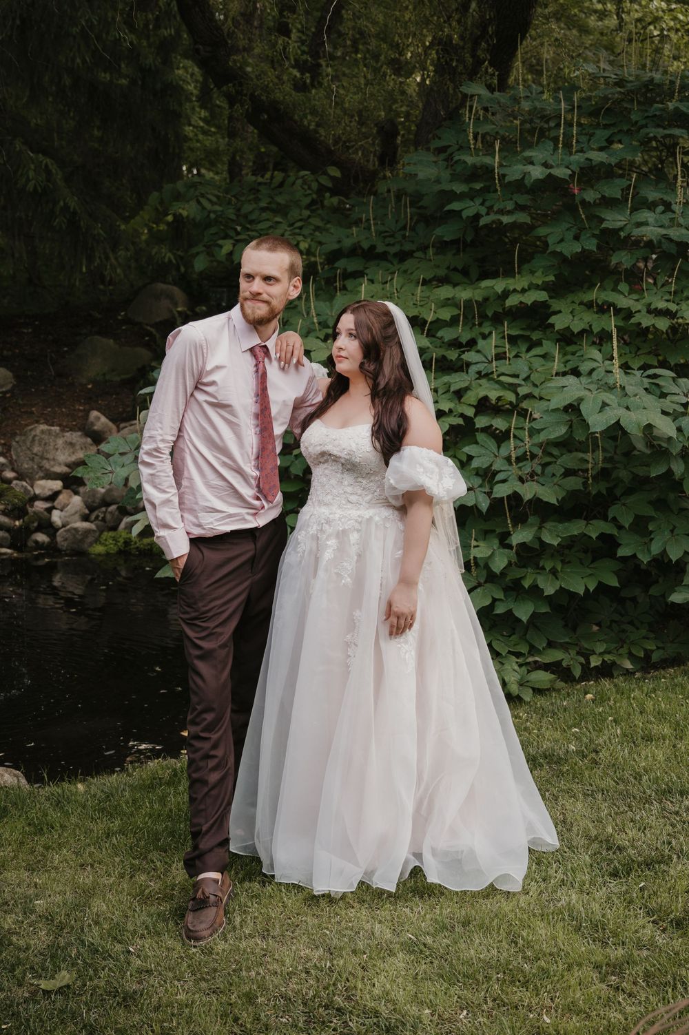 Couple poses outdoors near a creek, bride in off-shoulder tulle gown with veil, groom in pink shirt and burgundy tie.