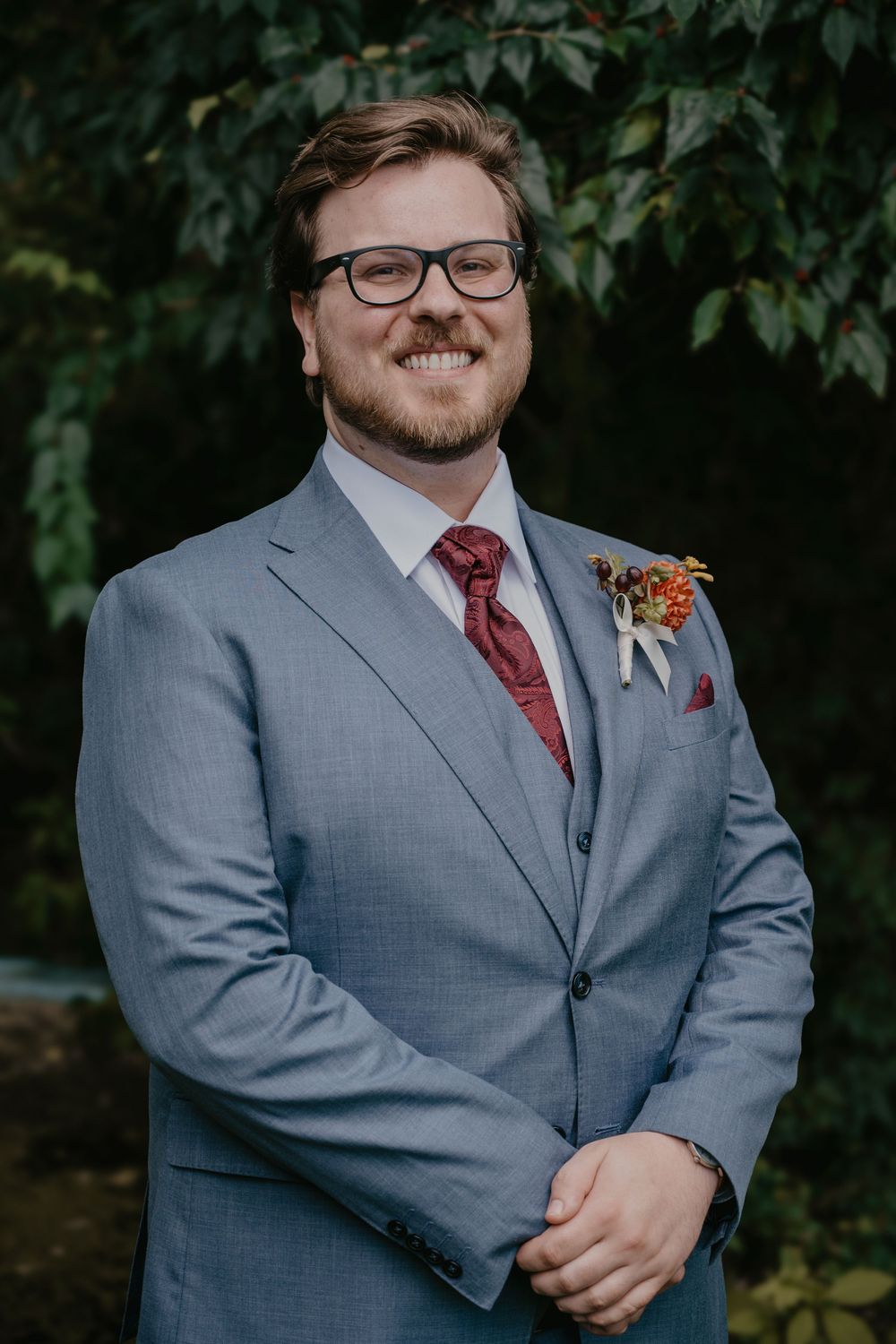 Smiling groom in light gray suit with burgundy tie and floral boutonniere outdoors.