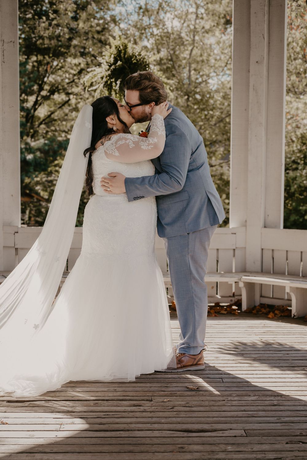 Bride and groom share first kiss in white gazebo with sunlight and flowing veil.