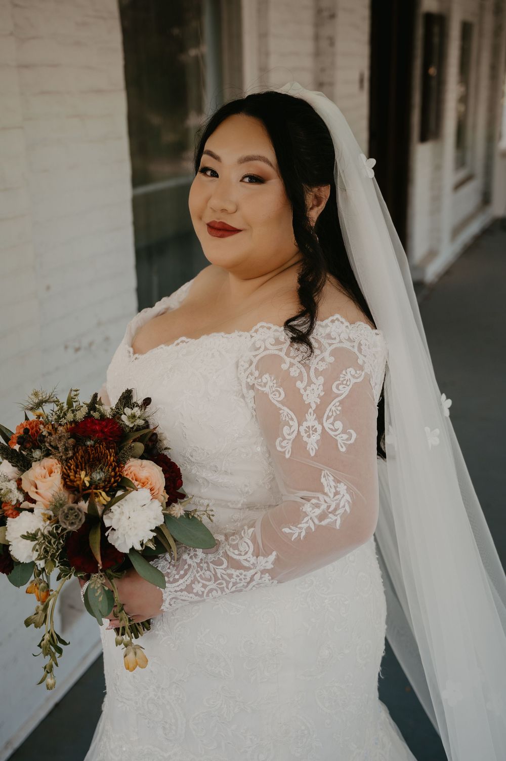 Bride in long-sleeve lace wedding gown holding autumn bouquet with veil and bold red lip.