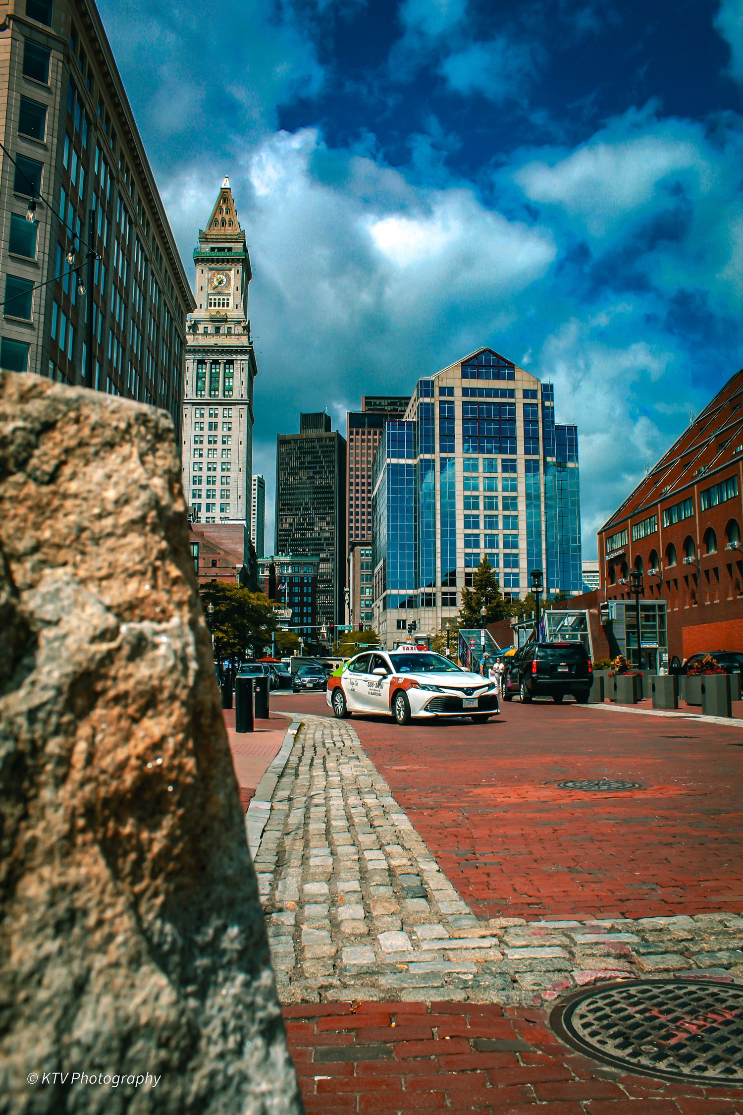 Boston Custom House clock tower rising above cobblestone street with modern glass skyscrapers nearby.
