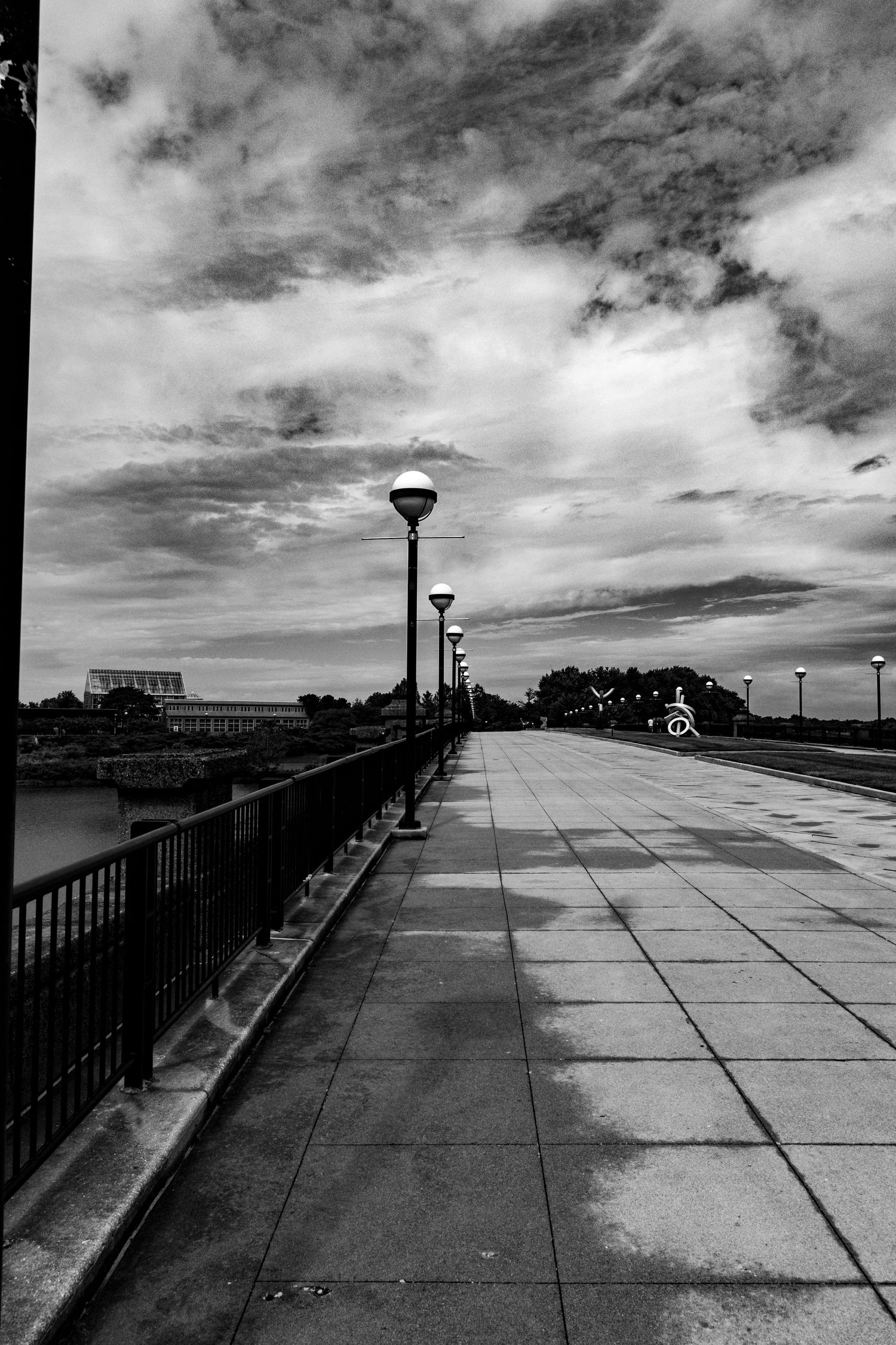 Black and white photo of an empty urban bridge walkway with lamp posts under dramatic cloudy sky.