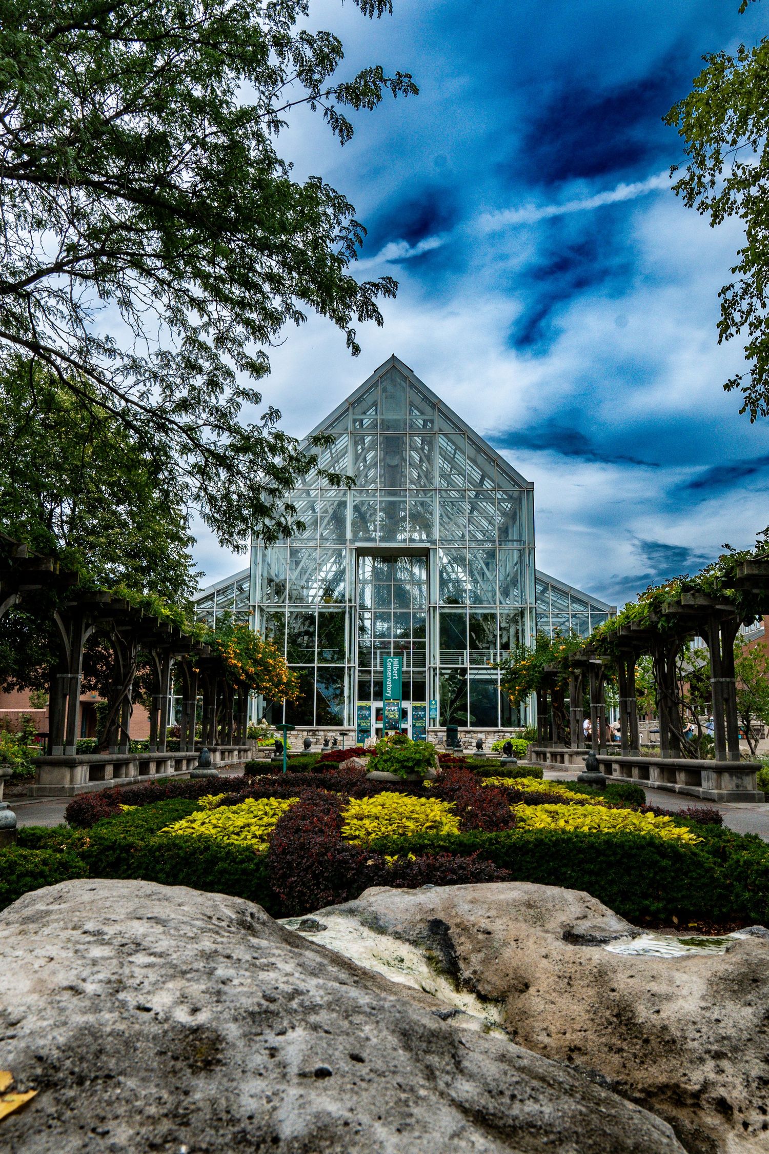 Beautiful glass greenhouse botanical garden with colorful flower beds and lush trees under dramatic sky.